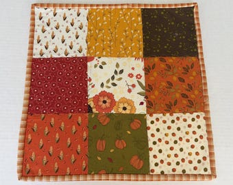 May include: A square fabric quilt featuring a nine-patch grid design with various autumn-themed patterns. The squares display motifs of corn, leaves, flowers, and pumpkins in shades of orange, yellow, brown, red, and green, finished with a plaid border.