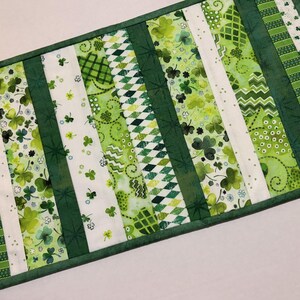 May include: A rectangular table runner featuring a patchwork design with various green fabrics. The design includes white stripes with shamrock patterns, green diamond patterns, and solid green stripes. The edges are trimmed with a solid green border.