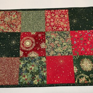 May include: A quilted placemat featuring a patchwork design with various Christmas-themed fabrics. The squares showcase patterns in red, green, and gold, including snowflakes, stars, and floral motifs. The edges are finished with a dark green border.