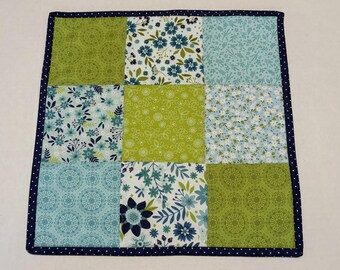 May include: A square patchwork quilt featuring a nine-block grid pattern. The fabric squares display various floral and geometric prints in shades of lime green, light blue, and dark navy. The entire piece is finished with a dark navy polka dot border.