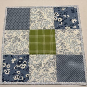 May include: A square quilted fabric mat featuring a nine-patch grid design. The pattern includes alternating squares of blue and white floral prints, a blue geometric diamond pattern, and a central square of solid green with white grid lines.