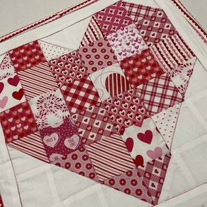May include: A white quilted heart-shaped wall hanging with a pink and red patchwork design. The patchwork includes various patterns of hearts, polka dots, stripes, and gingham. The text "Miss U" and "True Love" is visible on the patchwork.