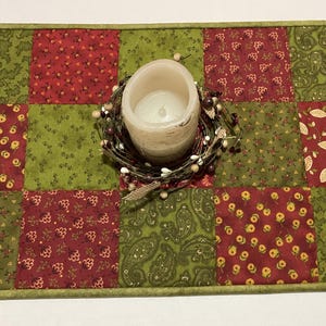 Puede incluir: Camino de mesa acolchado con un dise&ntilde;o de patchwork en rojo y verde. Una vela de color crema se encuentra encima del camino, rodeada por una peque&ntilde;a corona de bayas. El camino de mesa mide aproximadamente 30 cm de ancho.