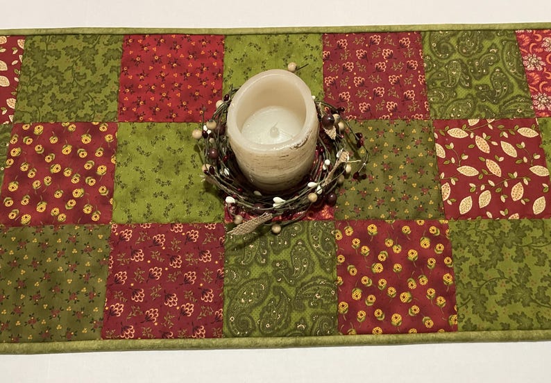 Puede incluir: Camino de mesa acolchado con un dise&ntilde;o de patchwork en rojo y verde. Una vela de color crema se encuentra encima del camino, rodeada por una peque&ntilde;a corona de bayas. El camino de mesa mide aproximadamente 30 cm de ancho.