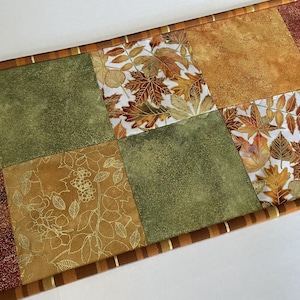 May include: A fall-themed table runner with a patchwork design. The runner features squares of various colors, including shades of green, gold, and red. Some squares have leaf patterns, while others have textured or floral designs. The edges are trimmed with a striped border.