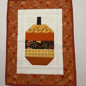 May include: A small quilted wall hanging featuring a patchwork acorn design in shades of orange, brown, and yellow. The acorn is surrounded by a white scalloped border and a brown outer border with a small floral pattern.
