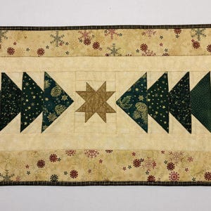 May include: A festive Christmas table runner featuring a central design of green triangle trees and a gold star. The runner is approximately 14 inches wide and has a beige background with snowflake and floral patterns.