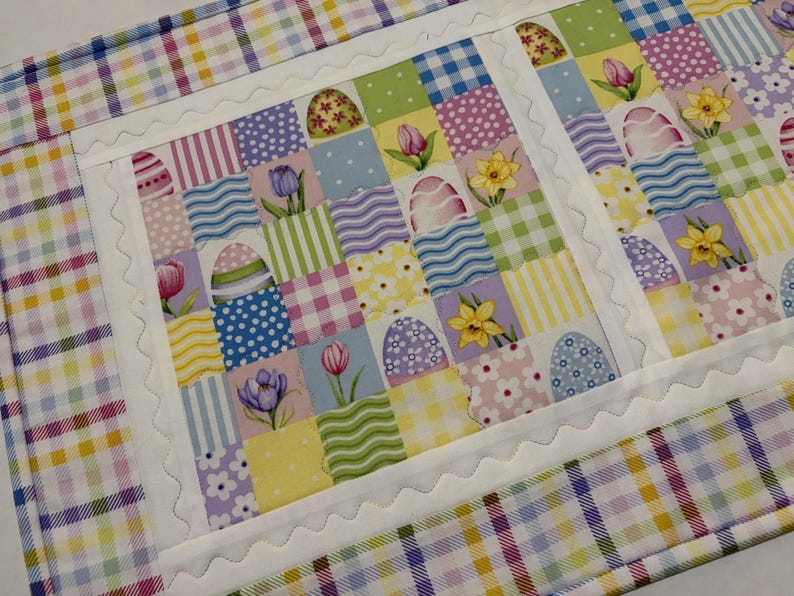 Pode incluir: Um caminho de mesa de P&aacute;scoa acolchoado com um design de patchwork. O centro apresenta quadrados com ovos de P&aacute;scoa, tulipas e narcisos. A borda &eacute; um padr&atilde;o xadrez colorido. O caminho de mesa tem cerca de 36 cm de largura e 102 cm de comprimento.