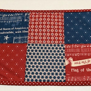 May include: A rectangular, patriotic placemat with a red, white, and blue patchwork design. The placemat features squares with star and floral patterns, musical notes, and the text "United States of America" and "Flag of the Free."