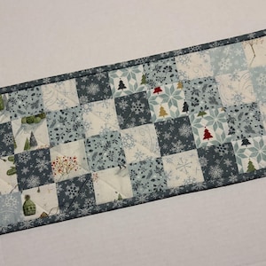 May include: A rectangular quilted table runner featuring a patchwork design of squares in shades of blue, white, and grey. The fabric patterns include snowflakes, small Christmas trees, winter foliage, and knit hats, all bordered by a dark grey trim.