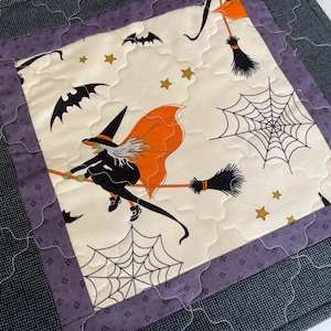 May include: A quilted Halloween placemat featuring a witch on a broomstick, bats, spiderwebs, and stars. The design is on a cream background with a purple and gray border. The placemat is square.