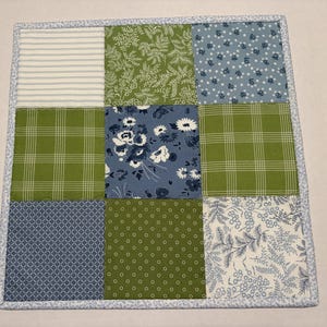 May include: A square quilted fabric mat featuring a nine-patch grid design. The squares display various patterns including green and white stripes, green foliage, blue floral prints, green plaid, and blue geometric shapes, all bordered by light blue trim.