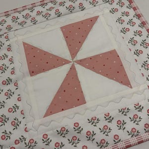 May include: A handmade quilted placemat with a pinwheel design. The center features a white square with four pink triangles dotted with darker pink circles. The border is white with small pink flowers and a red plaid trim.