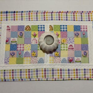 Pode incluir: Um jogo americano acolchoado com tema de P&aacute;scoa com um design de patchwork com ovos de P&aacute;scoa, flores e quadrados xadrez. Um casti&ccedil;al branco fica no centro. A borda &eacute; um padr&atilde;o xadrez colorido.