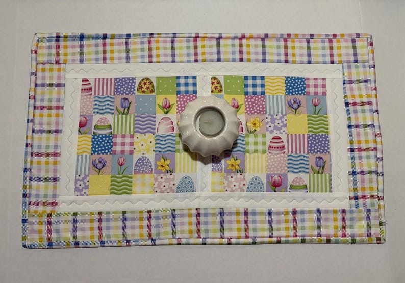 Pode incluir: Um jogo americano acolchoado com tema de P&aacute;scoa com um design de patchwork com ovos de P&aacute;scoa, flores e quadrados xadrez. Um casti&ccedil;al branco fica no centro. A borda &eacute; um padr&atilde;o xadrez colorido.