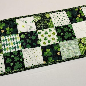 May include: A rectangular St. Patrick's Day table runner with a patchwork design. The squares feature various green patterns, including shamrocks, plaid, and diamond shapes, set against white and black backgrounds. The border is a dark green with small gold accents.