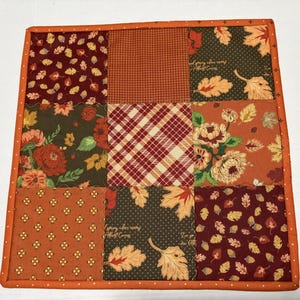 May include: A square fabric potholder featuring a nine-patch quilt design with various autumn-themed patterns including fall leaves, acorns, plaid, and floral prints in shades of orange, brown, red, and green, finished with an orange border.
