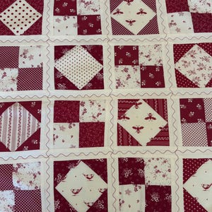 May include: A patchwork quilt featuring a repeating grid of squares in deep red and cream. The design includes alternating blocks of solid-colored squares, floral patterns, polka dots, stripes, and motifs of small bees and birds on cream backgrounds.