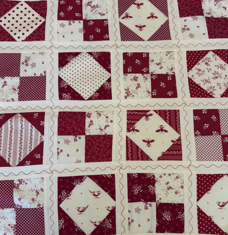 May include: A patchwork quilt featuring a repeating grid of squares in deep red and cream. The design includes alternating blocks of solid-colored squares, floral patterns, polka dots, stripes, and motifs of small bees and birds on cream backgrounds.
