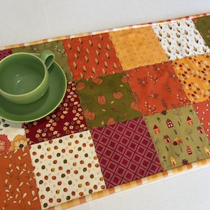 May include: A rectangular patchwork table runner featuring a variety of autumn-themed fabric squares in shades of orange, red, green, and white, with patterns including pumpkins, leaves, and houses, topped with a solid light green ceramic cup and saucer.