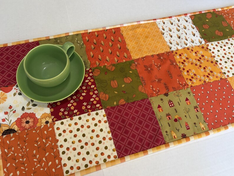 May include: A rectangular patchwork table runner featuring a variety of autumn-themed fabric squares in shades of orange, red, green, and white, with patterns including pumpkins, leaves, and houses, topped with a solid light green ceramic cup and saucer.