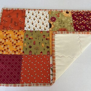 Peut inclure: Un set de table patchwork rectangulaire compos&eacute; d'une grille de neuf carr&eacute;s de tissu aux motifs automnaux dans des tons orange, jaune, rouge et vert. La bordure est en tissu &eacute;cossais et le dessous est en tissu uni couleur cr&egrave;me.