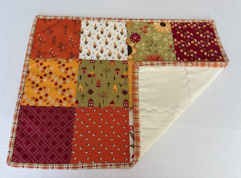 Peut inclure: Un set de table patchwork rectangulaire compos&eacute; d'une grille de neuf carr&eacute;s de tissu aux motifs automnaux dans des tons orange, jaune, rouge et vert. La bordure est en tissu &eacute;cossais et le dessous est en tissu uni couleur cr&egrave;me.