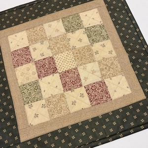 May include: A square quilted fabric mat featuring a central grid of sixteen patchwork squares in cream, olive green, and deep red with floral patterns. The center is framed by a textured tan border and a dark green outer border with small gold dots.