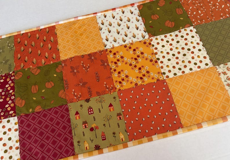 May include: A rectangular patchwork table runner featuring a variety of autumn-themed fabric squares in shades of orange, yellow, green, and burgundy. The patterns include pumpkins, corn, small houses, trees, flowers, and geometric diamond grids.