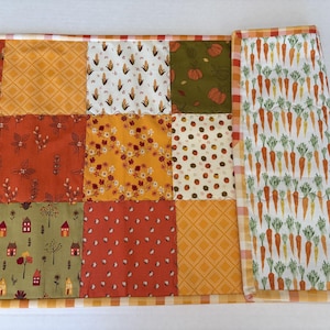 May include: A rectangular quilted table runner featuring a patchwork design of various autumn-themed fabrics including corn, pumpkins, small houses, and floral patterns, finished with a white and orange checkered border and a carrot print section.