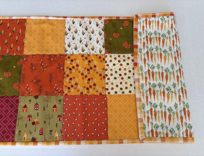 May include: A rectangular quilted table runner featuring a patchwork design of various autumn-themed fabrics including corn, pumpkins, small houses, and floral patterns, finished with a white and orange checkered border and a carrot print section.
