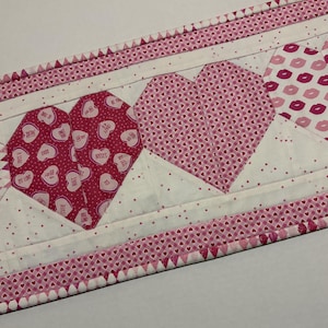 May include: A quilted table runner featuring a repeating pattern of pink and white hearts. The hearts are filled with various designs, including candy hearts and lip prints. The edges are trimmed with a pink heart pattern.