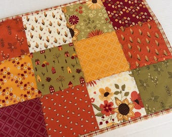 May include: A rectangular quilted fabric mat featuring a patchwork design of various squares in autumn colors including orange, yellow, olive green, and deep red. The patterns include sunflowers, corn stalks, small houses, floral prints, and geometric grids.