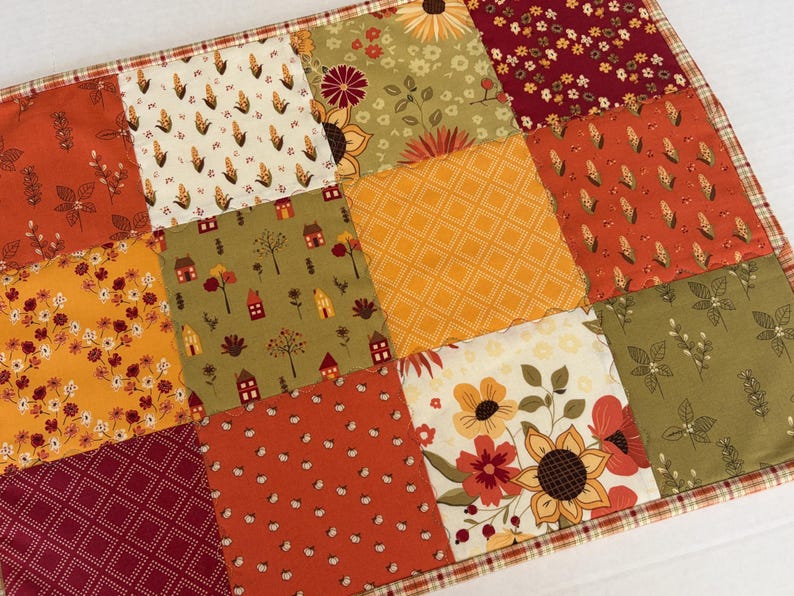 Peut inclure: Un tapis en tissu matelass&eacute; rectangulaire pr&eacute;sentant un motif en patchwork compos&eacute; de divers carr&eacute;s aux couleurs automnales, notamment orange, jaune, vert olive et rouge fonc&eacute;. Les motifs incluent des tournesols, du ma&iuml;s, des maisons et des fleurs.