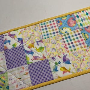 May include: A colorful patchwork table runner with a yellow border. The runner features a variety of Easter-themed fabrics, including bunnies, eggs, and gingham patterns. The text "Happy Hoppy Easter" is visible on the runner.