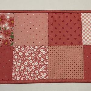 May include: A rectangular, quilted placemat with a patchwork design. The design features squares of various pink fabrics, including floral, polka dot, heart, and check patterns. The edges are finished with a solid pink binding.
