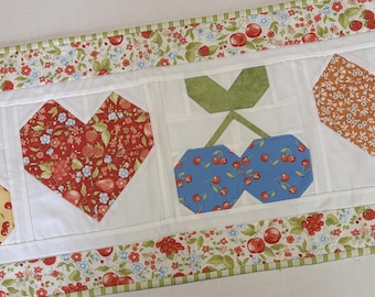 May include: A rectangular fabric table runner featuring a patchwork design with alternating blocks of cherry and heart motifs. The border displays a floral pattern with red cherries and green leaves on a white background, framing the central quilted squares.