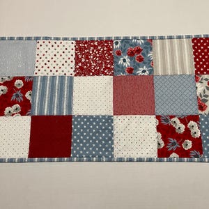 May include: A patchwork table runner with a red, white, and blue color scheme. The runner is made of various fabrics with different patterns, including floral, polka dot, and striped.