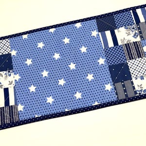 May include: A rectangular quilted table runner featuring a patchwork design in shades of blue and white. The center section has a blue background with white stars. The patchwork includes floral, striped, and checkered patterns. The runner is bordered with a dark blue dotted fabric.