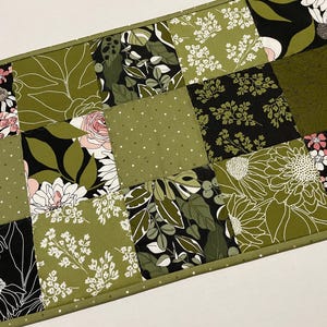 May include: A rectangular patchwork table runner featuring a variety of floral and leaf patterns in shades of green, black, and white. The design includes daisies, roses, and other botanical elements. The runner is bordered with a green trim.