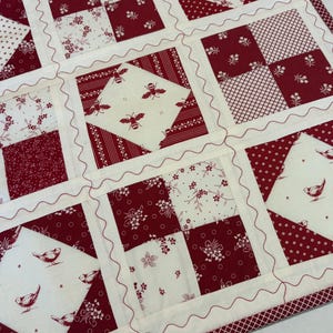 May include: A patchwork quilt featuring a repeating grid of squares in deep red and cream. The design includes alternating blocks of floral, striped, and polka dot patterns, alongside squares printed with small birds and bees, finished with red wavy stitching.