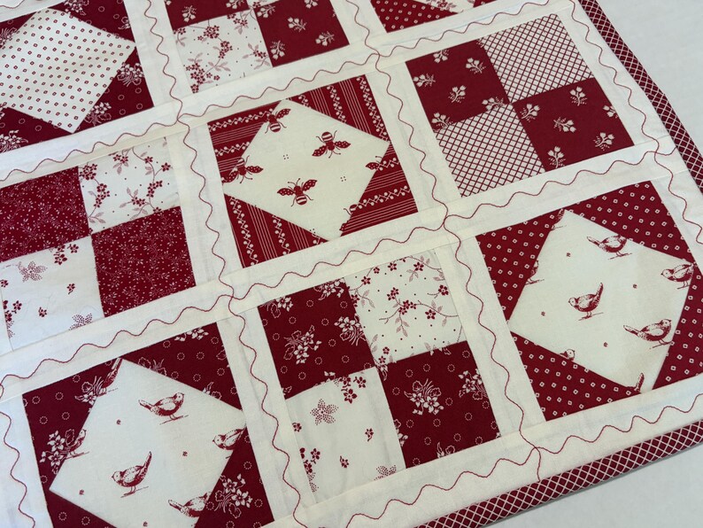 May include: A patchwork quilt featuring a repeating grid of squares in deep red and cream. The design includes alternating blocks of floral, striped, and polka dot patterns, alongside squares printed with small birds and bees, finished with red wavy stitching.