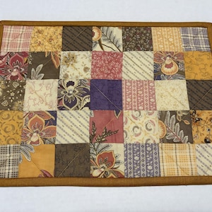 May include: A rectangular quilted fabric mat featuring a patchwork design of various square fabric pieces in shades of brown, gold, cream, purple, and red. The squares display floral, striped, and geometric patterns, all bordered by a solid brown trim.