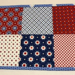 May include: A rectangular patchwork placemat with a blue border. The patchwork design features red, white, and blue fabrics with floral, star, and gingham patterns. The border is a solid blue with white polka dots and white stars.