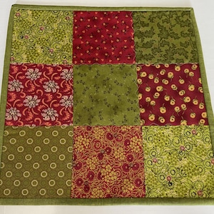 May include: A square, quilted patchwork item with nine fabric squares in shades of red and green. The squares feature floral and geometric patterns. A green border frames the piece. This item could be a trivet, placemat, or decorative accent.