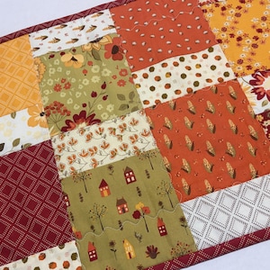 May include: A rectangular patchwork table runner featuring a variety of autumn-themed fabric squares. Patterns include yellow sunflowers, orange corn cobs, small houses with trees, and geometric diamond prints in shades of red, gold, and olive green.