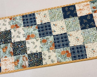 May include: A rectangular patchwork table runner featuring a grid of squares with various patterns including navy blue plaid, cream floral prints, orange pumpkins, and green foliage, all bordered by a thin yellow trim with a repeating pattern.