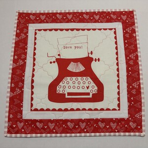 May include: A square, quilted fabric with a red and white heart pattern border. The center features a red typewriter illustration with the words "love you!" on a paper. The quilt is approximately 12 inches square.