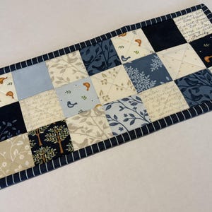May include: A rectangular quilted table runner featuring a patchwork design of squares in shades of navy blue, light blue, and cream. The fabric squares display patterns of birds, trees, leaves, and cursive script text, finished with a navy striped border.