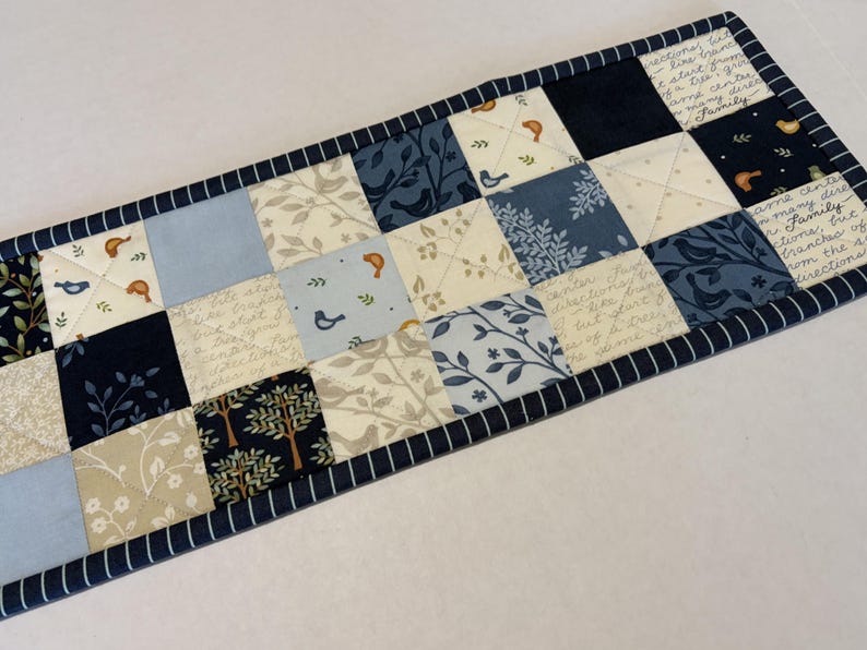 May include: A rectangular quilted table runner featuring a patchwork design of squares in shades of navy blue, light blue, and cream. The fabric squares display patterns of birds, trees, leaves, and cursive script text, finished with a navy striped border.